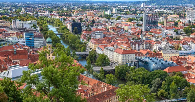 A panoramic view of Graz, Austria, showcasing red-roofed buildings, the Mur River cutting through the city, and lush greenery. Notable structures include the Kunsthaus Graz with its distinctive modern architecture. The surrounding hills and skyline complete the scenic vista.