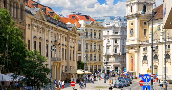 A bustling street scene in Vienna features historic, ornate buildings with architectural details. The prominent clock tower of St. Michael's Church is visible. People walk along the street, and various signs and outdoor seating areas are present. The sky is partly cloudy.