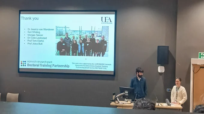 Alex and Emma standing near a podium in a lecture hall. The screen behind them displays a "Thank you" slide with the University of East Anglia logo. The slide lists acknowledgments for several individuals, including Dr. Jessica van Wonderen and Prof. Julea Butt, and displays a group photograph of a research team outdoors. The bottom of the slide mentions the Norwich Research Park Doctoral Training Partnership.