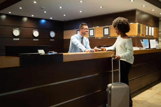 A person checking into a hotel, interacting with a receptionist at the front desk. The lobby features elegant wood paneling and multiple clocks displaying various times.