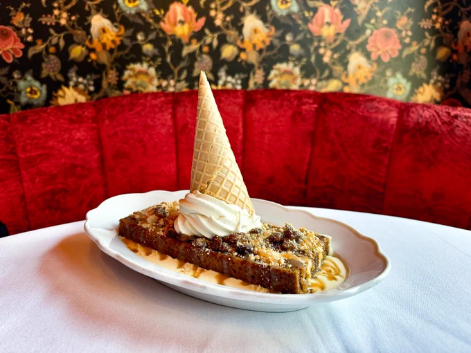Slice of nutty cake topped with whipped cream and an upside-down ice cream cone on a white plate against a red velvet booth and floral wallpaper.