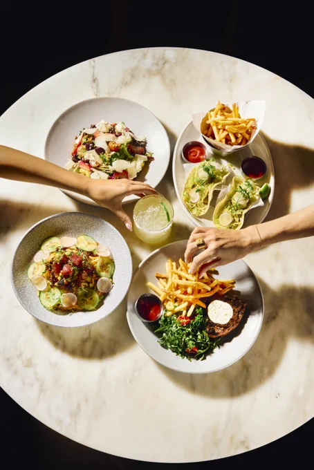 Overhead view of a marble table with plates of tacos, fries, salad, and a pasta dish, two hands reaching for food and a drink.