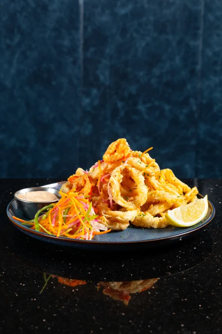 Crispy golden fried calamari rings served with a lemon wedge, colorful shredded vegetable garnish, and a side of creamy dipping sauce on a dark plate.
