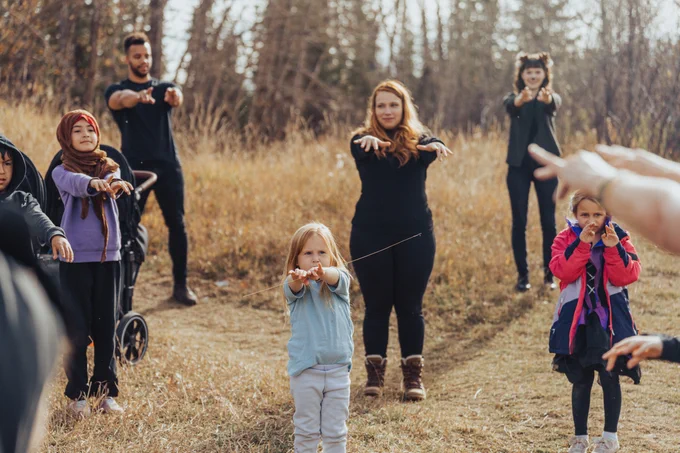 IMAGE: Outdoors photo showing a diverse group of adults and children standing in a tall-grass field near a forest. They are participating in a hands-on activity organized by the southern Alberta chapter of the Canadian Parks and Wilderness Society (CPAWS). (Photo Credit: CPAWS/Amber Toner)