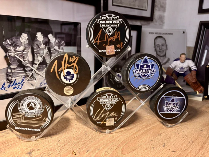 A perspect puck display with 6 pucks sits on a desk in front of framed photos of the maple leafs