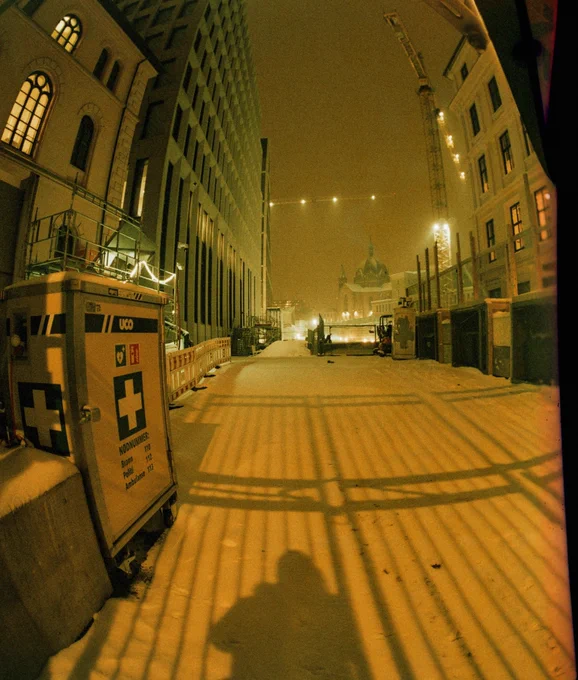 Entrance to a snow covered building site in the city at night. A church and a tower crane in the distance. Shadow of the photographer and a fence on the snow.