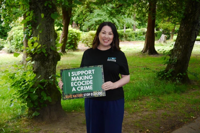 Photo of Monica Lennon in a park next to trees and grass holding a poster that says I support making ecocide a crime, save nature, stop ecocide, wearing a stop ecocide international t-shirt.