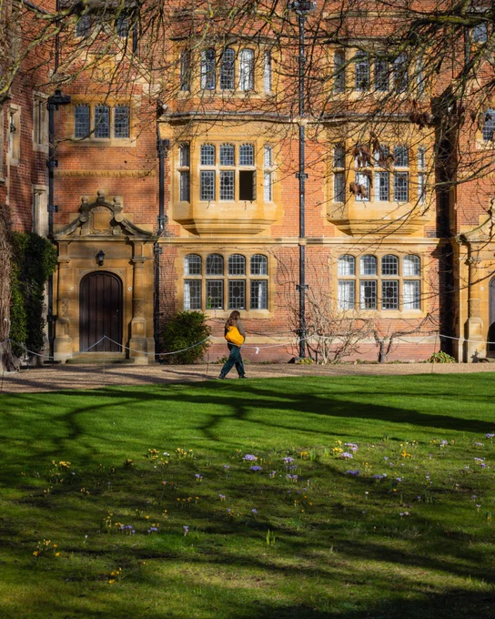 The image shows a girl with a yellow bag walking across a path at Trinity hall in Cambridge. There is green grass with some spring flowers at the bottom of the image.