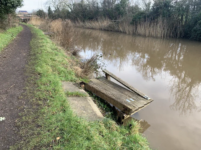 A 'before' picture of a weathered, rotten and partially broken wooden platform protruding into a river from the bank. A footpath runs alongside the bank. The river is brown and calm, reflecting trees lining the opposite bank. Picture taken in January 2025.