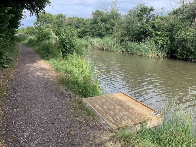 The 'after' picture of a new wooden fishing platform protruding into the river from the bank. A footpath runs alongside the riverbank. The river is calm and reflects a cloudless sky. Vegetation runs alongside both riverbanks. Picture taken in May 2025.