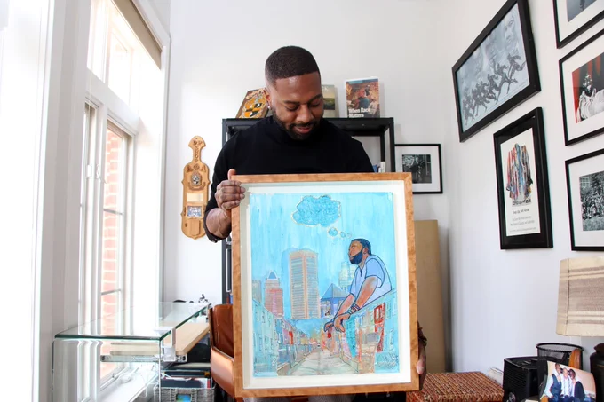 A Black man gazes at a framed image he is holding. He wears a black top.