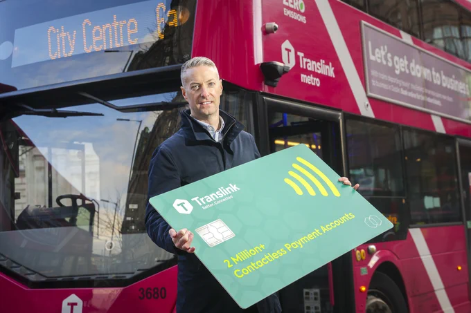A male adult standing in front of a pink Translink Metro bus while holding a large promotional contactless payment card.