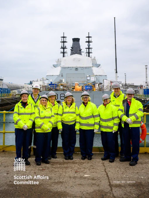 Members of the Scottish Affairs Committee in high vis jackets standing in front of a large warship