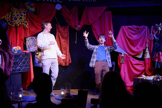 Photograph of two people onstage. One laughs heartily at their fellow performer. The second person has wide open eyes, stands with their knees slightly bent, and their arms over their head with the fingers slightly curled. They are possibly demonstrating something explosive.