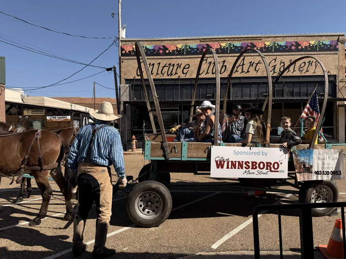 A cowboy with his back to the camera looks at a wagon carrying people past a gallery storefront.