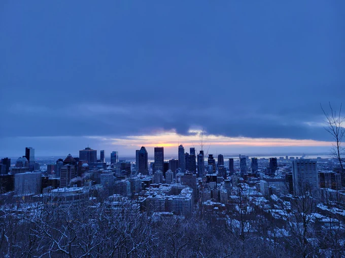 Sunrise breaks through dark grey clouds cover, looking out over Montreal from the overlooking on Mont Royal. There are some bare sumac branches in the foreground, then the city scape.