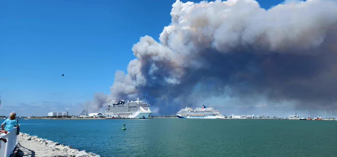 Thick smoke billowing over a harbor with two large cruise ships docked under a clear blue sky.