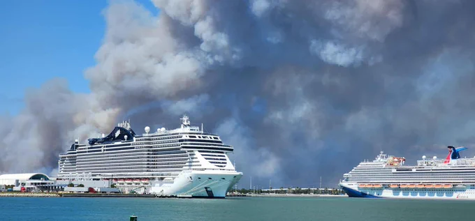 Two large cruise ships docked near a port with thick dark smoke billowing in the sky behind them.