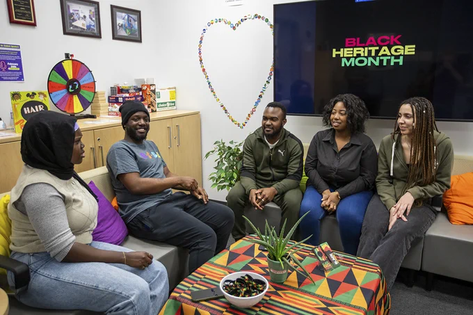 A group of people are sitting on two couches. A screen behind them reads Black Heritage Month.
