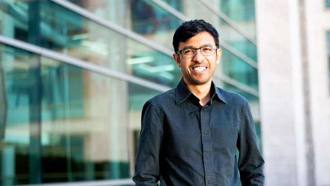 Smiling man with glasses and short black hair standing in front of a glass and stone building wearing a dark collard shirt.