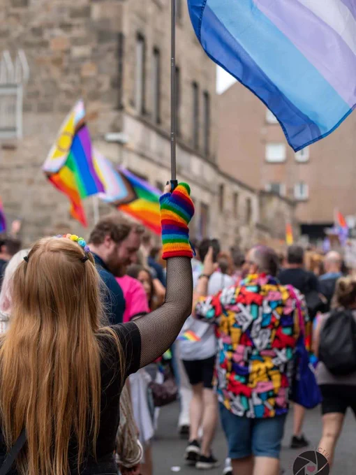 Person wearing rainbow gloves holding a transgender pride flag at a crowded outdoor LGBTQ  event.