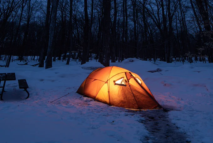 A glowing tent illuminated from within is pitched on snowy ground in a forest at twilight.