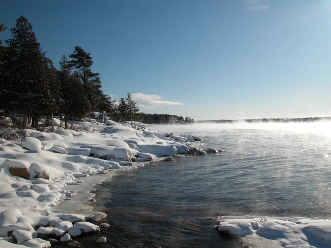 Winter scene at a snowy lakeshore with mist rising from the lake under a clear blue sky.