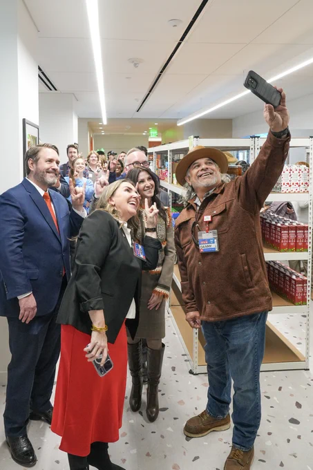 A group of people in the UT Outpost are gathered for a selfie. One individual in a hat holds the phone up to capture the moment. Smiling faces can be seen among the group, dressed in a mix of formal and casual attire. 