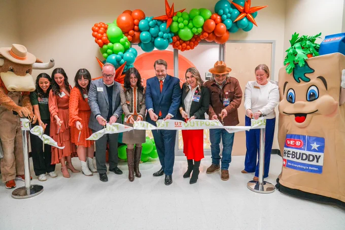 A group of people in formal attire are participating in a ribbon-cutting ceremony for the UT Outpost, flanked by colorful balloon decorations. Two mascots, Hook 'Em and H-E-Buddy, are also cutting the ribbon. 