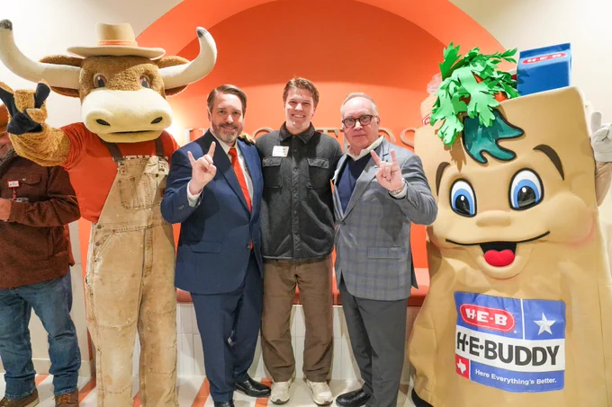 A group of people and mascots stand in front of an orange and white backdrop. Notable figures include the Texas Longhorns mascot and the H-E-Buddy mascot from H-E-B, both holding up hand signs. Two individuals in suits and one person in casual attire are also part of the group.