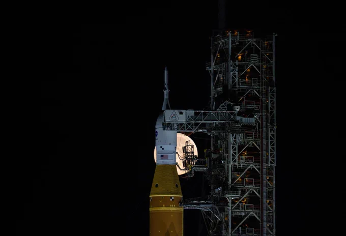 Image shows A full Moon is seen shining behind NASA’s SLS (Space Launch System) and Orion spacecraft, atop the mobile launcher in the early hours of Feb. 1, 2026. The rocket is currently at Launch Pad 39B at NASA’s Kennedy Space Center in Florida, as teams are preparing for a wet dress rehearsal to practice timelines and procedures for the launch of Artemis II. Photo credit: NASA/Sam Lott