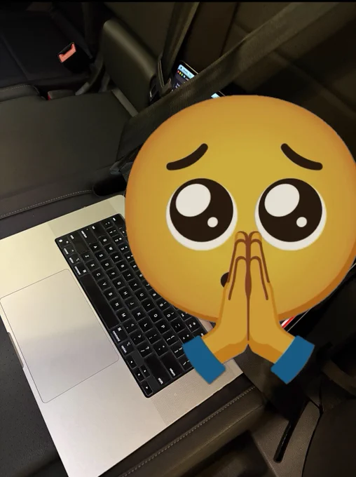 A MacBook Pro sitting on a back seat of an SUV, opened, and buckled in. The screen is masked by a pleading eyes emoji with praying hands over the mouth.