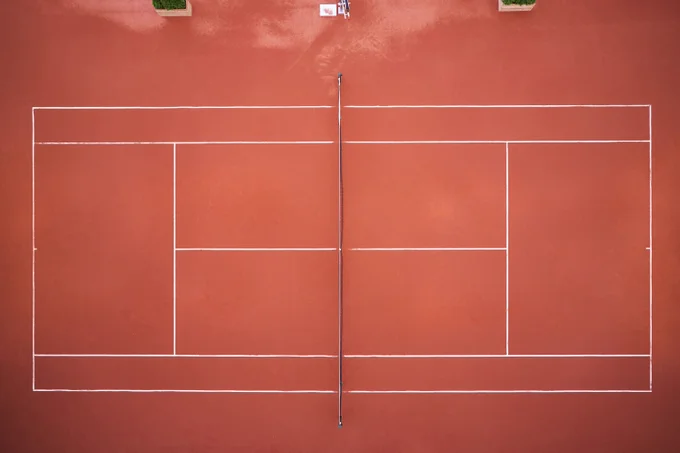 Aerial view of an empty red clay tennis court with white boundary lines and a central net.