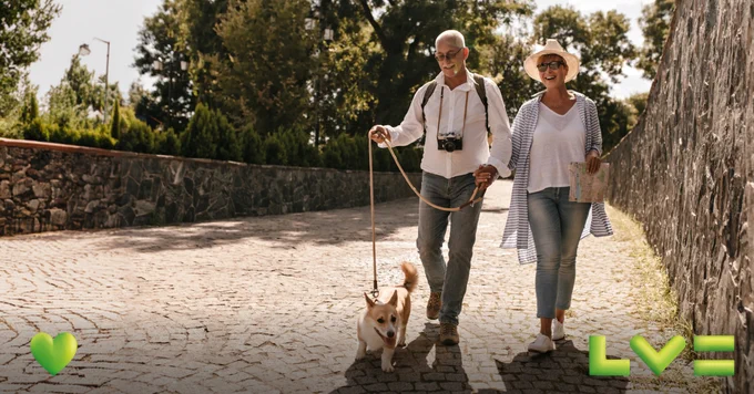 An older couple are enjoying themselves, walking a dog along a paved path on a sunny day. One holds a dog leash and a camera, while the other holds a map and wears a sunhat. A green heart and the LV logo are at the bottom of the image.