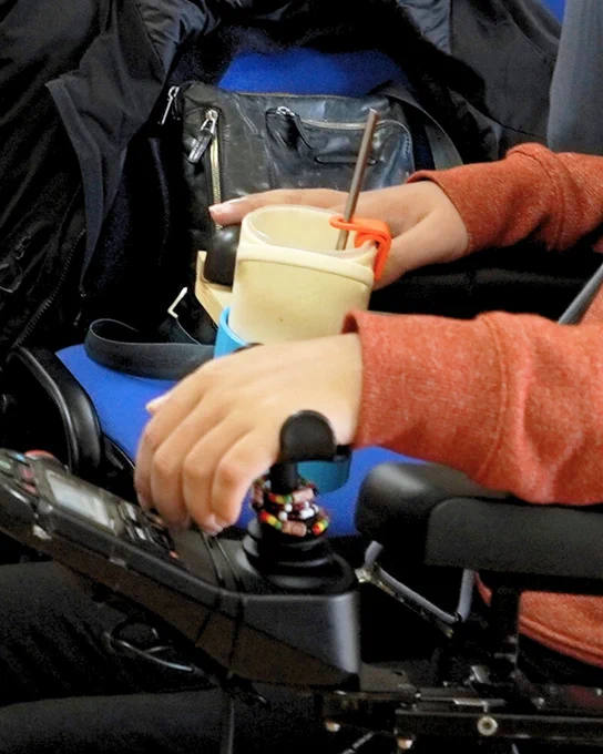 Detalle de la mano de una persona sobre el mando de una silla de ruedas junto a un vaso con soporte para pajita impreso en 3D.