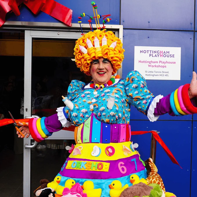 A panto dame dressed in colourful 'nanny' costume cuts a red ribbon on a blue building which says Nottingham Playhouse Workshops. They smile broadly at the camera and open their arms wide.