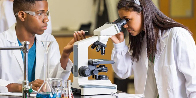 Two people in lab coats are examining a sample with a microscope in a laboratory. Several beakers containing colorful liquids are on the table.