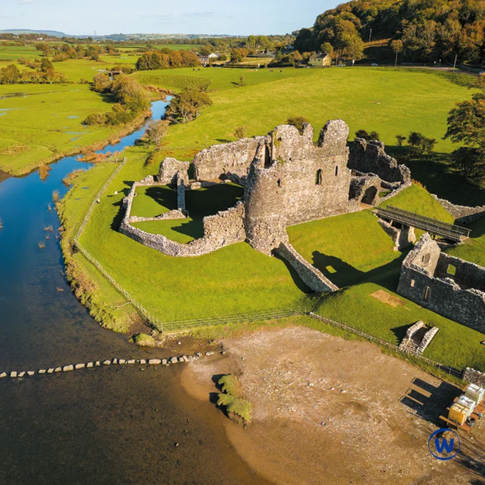 The photo is of the River Ewenny in Wales, surrounded by green fields and trees. The blue Ofwat logo, a W in a circle, is in the bottom right hand corner of the photo.