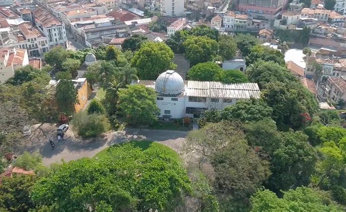 Fotografia aérea do prédio do Observatório do Valongo da UFRJ. No centro da imagem, em destaque, a edificação, de cor clara, tem uma cúpula hemisférica característica de observatórios astronômicos, utilizada para abrigar equipamentos de observação. Ao redor do observatório, há jardins e áreas arborizadas com caminhos internos, criando contraste com o ambiente urbano visível na parte superior da imagem.