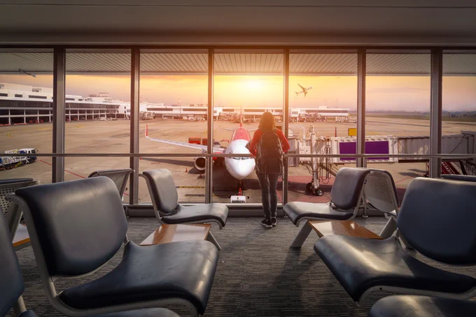 Person with a backpack standing at an airport gate, looking out at an airplane during sunset.