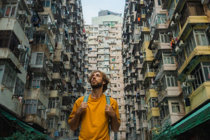 Man with a backpack standing between tall apartment buildings in Hong Kong, looking up.