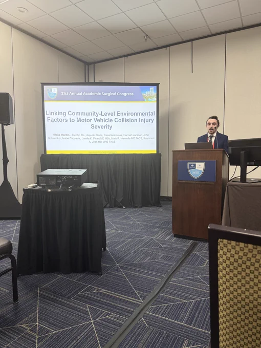 Blake Hardin, cis-white man, dressed in a suit at a podium presenting his research abstract titled “Linking Community-Level Environmental Factors to Motor Vehicle Collision Injury Severity”