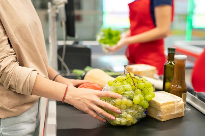 At a checkout line, a person in a beige shirt places bagged grapes on the conveyor belt next to a red pepper and butternut squash. A cashier in a red apron holds a head of lettuce. There are bottles of oil and packaged bread nearby.