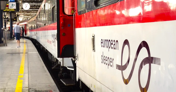 The image shows an "European Sleeper" train at a railway platform. The train has a red and white exterior, and a person walks nearby. Clocks and signage are visible above.