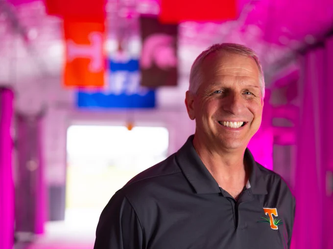 John Sorochan smiling in a brightly lit hallway with colorful banners in the background, including one with the Tennessee Volunteers logo.