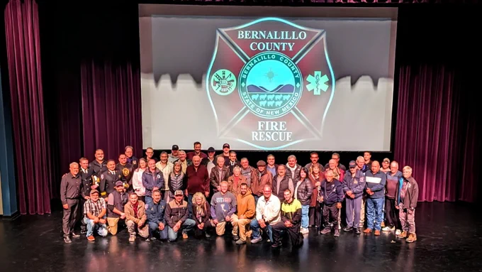 A group of people is gathered onstage in a theater, posing for a photo. Behind them is a large screen displaying the logo for Bernalillo County Fire Rescue. The stage curtains are visible on the sides.