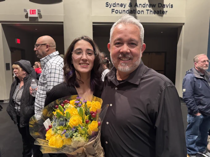 A person holding a bouquet of colorful flowers stands next to another person inside the Sydney & Andrew Davis Foundation Theater. Several people are visible in the background, and the setting appears to be a public event.