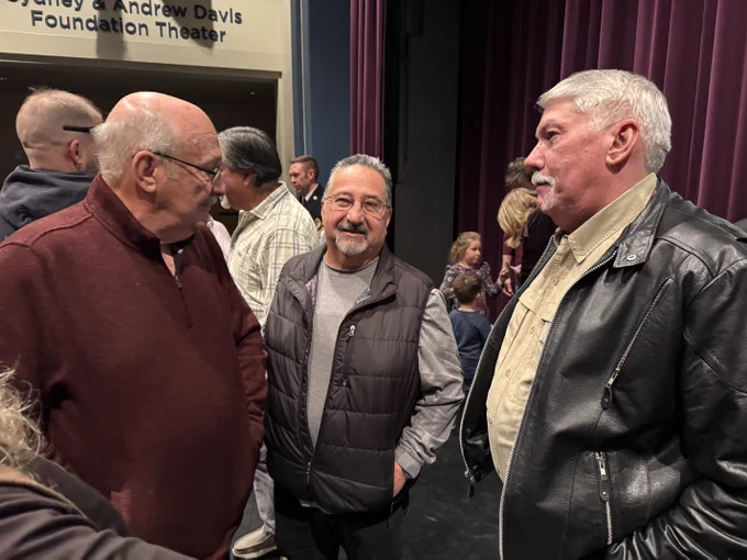 Three people conversing in a theater lobby. The background features a sign that reads "Charline & Sydney & Andrew Davis Foundation Theater". Other attendees are visible in the background.