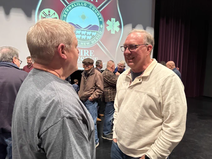 A group of people stands in a theater-like setting, engaging in conversation. Two individuals in the foreground are talking face-to-face. In the background, a projected image displays the Bernalillo County Fire Rescue logo on a screen.