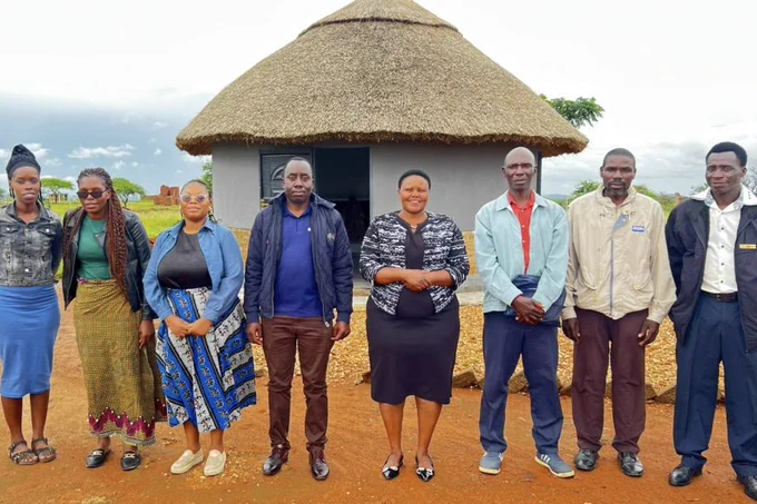 Eight people stand together side-by-side in front of a small building in rural Zimbabwe.